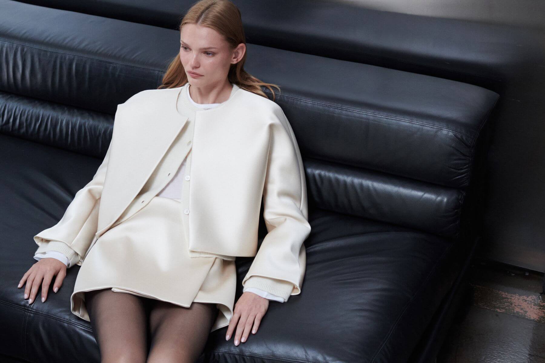 Model is sitting on black couch in champagne colored bobbi jacket and skirt.