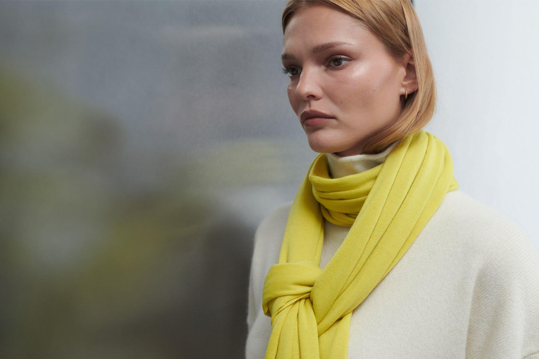Close up of model with yellow scarf over creme turtleneck sweater, looking to the side.