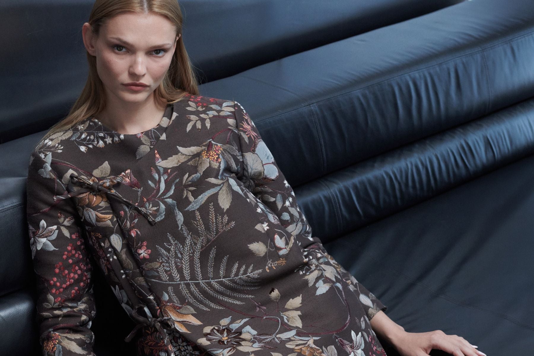 Model is leaning down on couch in brown floral jacket.