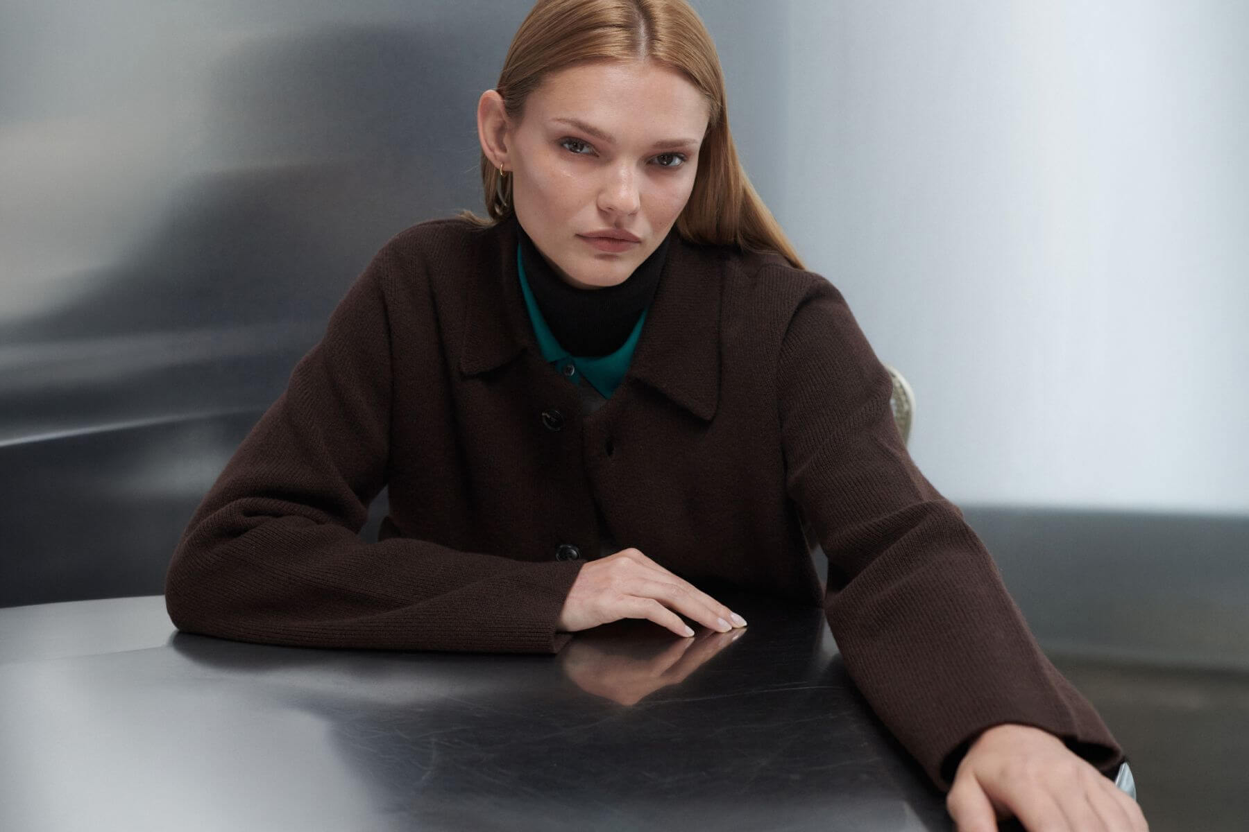 Model is sitting down with arms on table wearing chocolate zoe coat layered over teal sweater and turtleneck.