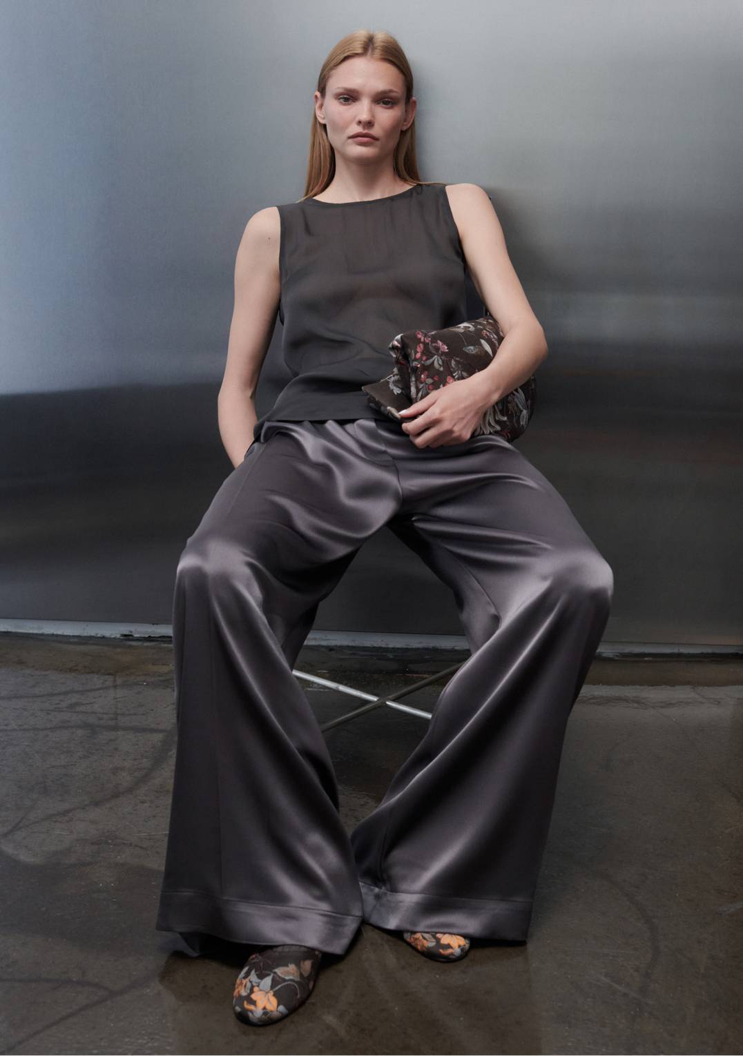 Model is leaning down in chair in farrah sleeveless top with grey satin pants.