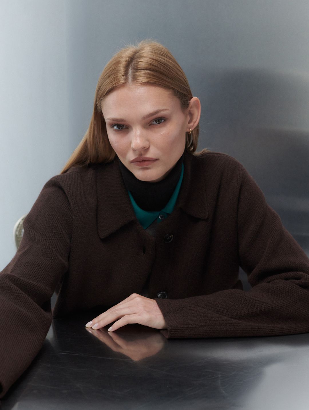 Model is leaning down on table in zoe chocolate sweater jacket layered over teal collar and turtleneck.